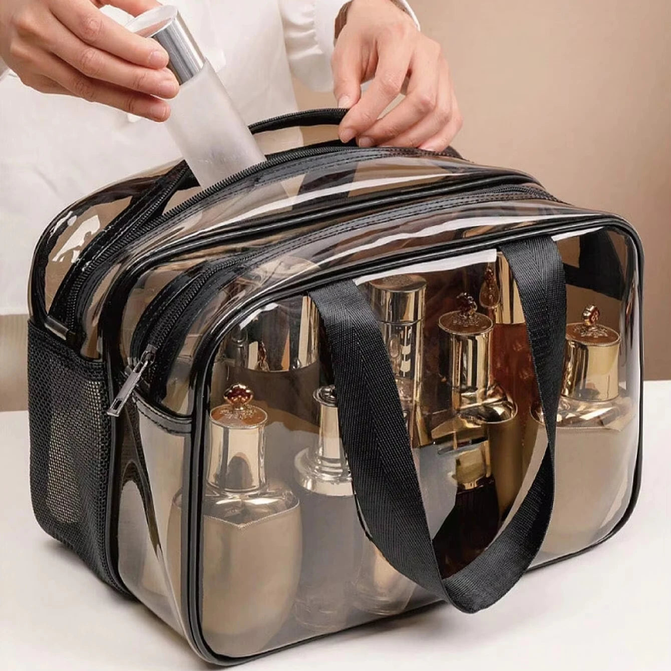 Une trousse de toilette femme transparente avec plusieurs compartiments zippés. À l'intérieur, on aperçoit divers produits de beauté et flacons de cosmétiques. Une main est en train de placer un flacon supplémentaire dans l'un des compartiments. La trousse semble pratique pour organiser et transporter les affaires de toilette. Un encart montre également la trousse remplie et ouverte, offrant une bonne visibilité du contenu.