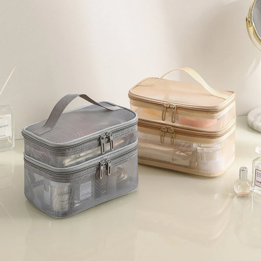Trousse de toilette femme transparente à double compartiment, l'une grise et l'autre beige, posées sur une surface claire.
Chaque trousse possède deux zips argentés et une poignée de transport sur le dessus.
Des produits de beauté et de soin sont visibles à travers le matériau transparent.