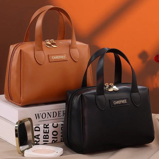 Trousse de toilette femme : Deux élégantes trousses de toilette en cuir, l'une brune et l'autre noire, sont posées sur des livres, avec un miroir de maquillage doré à proximité. Elles arborent une fermeture éclair dorée et une petite plaque rectangulaire portant l'inscription "CAREFREE". L'arrière-plan est un dégradé de tons chauds, suggérant une ambiance luxueuse.