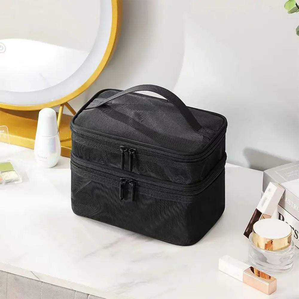 Une trousse de toilette noire en maille a une poignée sur le dessus et deux fermetures éclair sur le dessus, assise sur un comptoir en marbre blanc. À côté de la trousse se trouvent divers produits de beauté, notamment un flacon blanc, un pot en verre et des cosmétiques dorés et blancs. À l'arrière-plan flou, on aperçoit un miroir rond avec un éclairage doré. Le nom à afficher est 