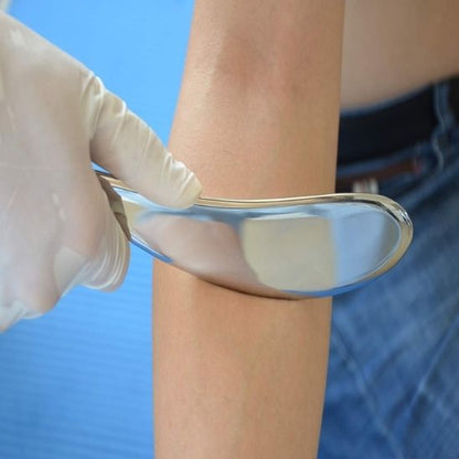 Une main gantée de blanc utilise un outil métallique lisse, probablement un instrument de gua sha, sur l'avant-bras d'une personne. L'outil reflète légèrement la lumière. L'arrière-plan est un drap bleu clair, et une partie d'un pantalon en jean est visible sur le côté droit. La scène suggère un traitement de bien-être ou une thérapie manuelle.