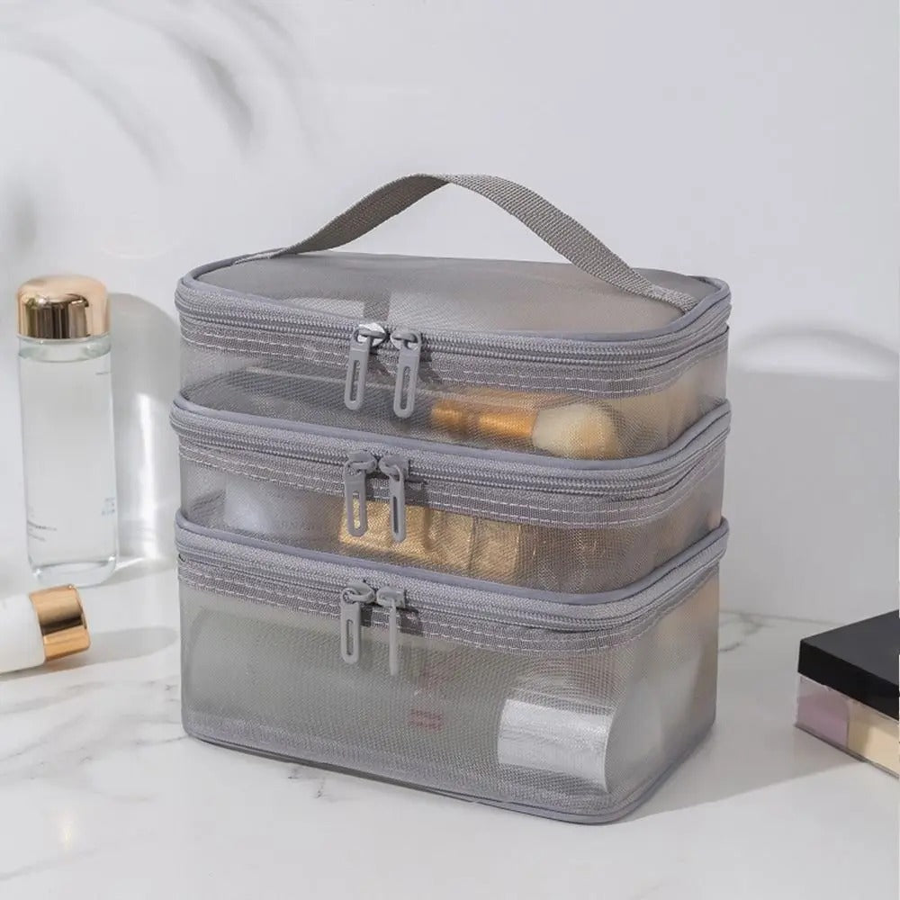 Trois trousses de toilette femme empilées en maille grise semi-transparente, chacune avec une fermeture éclair argentée et une petite poignée.
La trousse supérieure contient des pinceaux de maquillage aux manches blancs et aux poils beiges.
La trousse du milieu renferme divers produits de beauté aux emballages dorés.
La trousse inférieure est partiellement remplie de flacons et de tubes blancs.