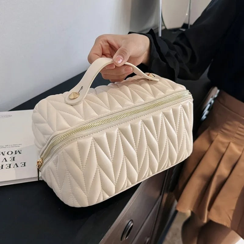 Une main tenant une Trousse de toilette femme blanche et élégante, avec un motif matelassé en losanges.
La trousse est dotée d'une fermeture éclair dorée et d'une poignée sur le dessus, accentuée par des détails dorés.
À l'arrière-plan flou, on aperçoit une personne portant une jupe marron et un vêtement noir, devant un meuble sombre.