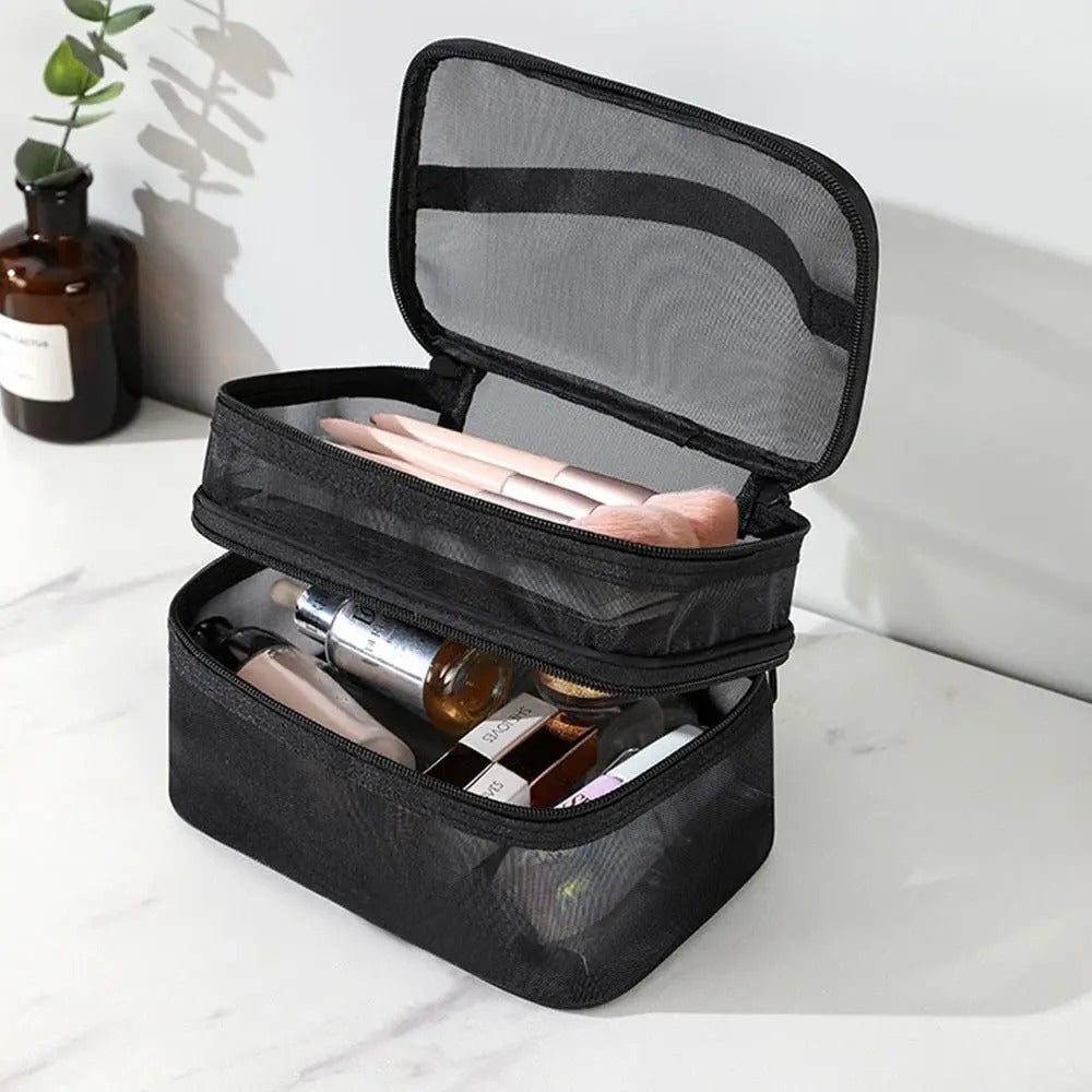 Une trousse de toilette femme noire en maille à double compartiment est ouverte sur un comptoir en marbre blanc. Le compartiment supérieur contient des pinceaux de maquillage aux manches roses. Le compartiment inférieur contient divers produits de beauté, notamment des tubes et des flacons. Un vase en verre brun avec une plante verte est visible à l'arrière-plan.