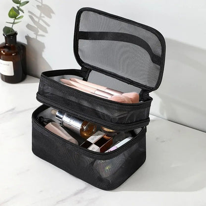 Une trousse de toilette femme noire en maille à double compartiment est ouverte sur un comptoir en marbre blanc. Le compartiment supérieur contient des pinceaux de maquillage aux manches roses. Le compartiment inférieur contient divers produits de beauté, notamment des tubes et des flacons. Un vase en verre brun avec une plante verte est visible à l'arrière-plan.