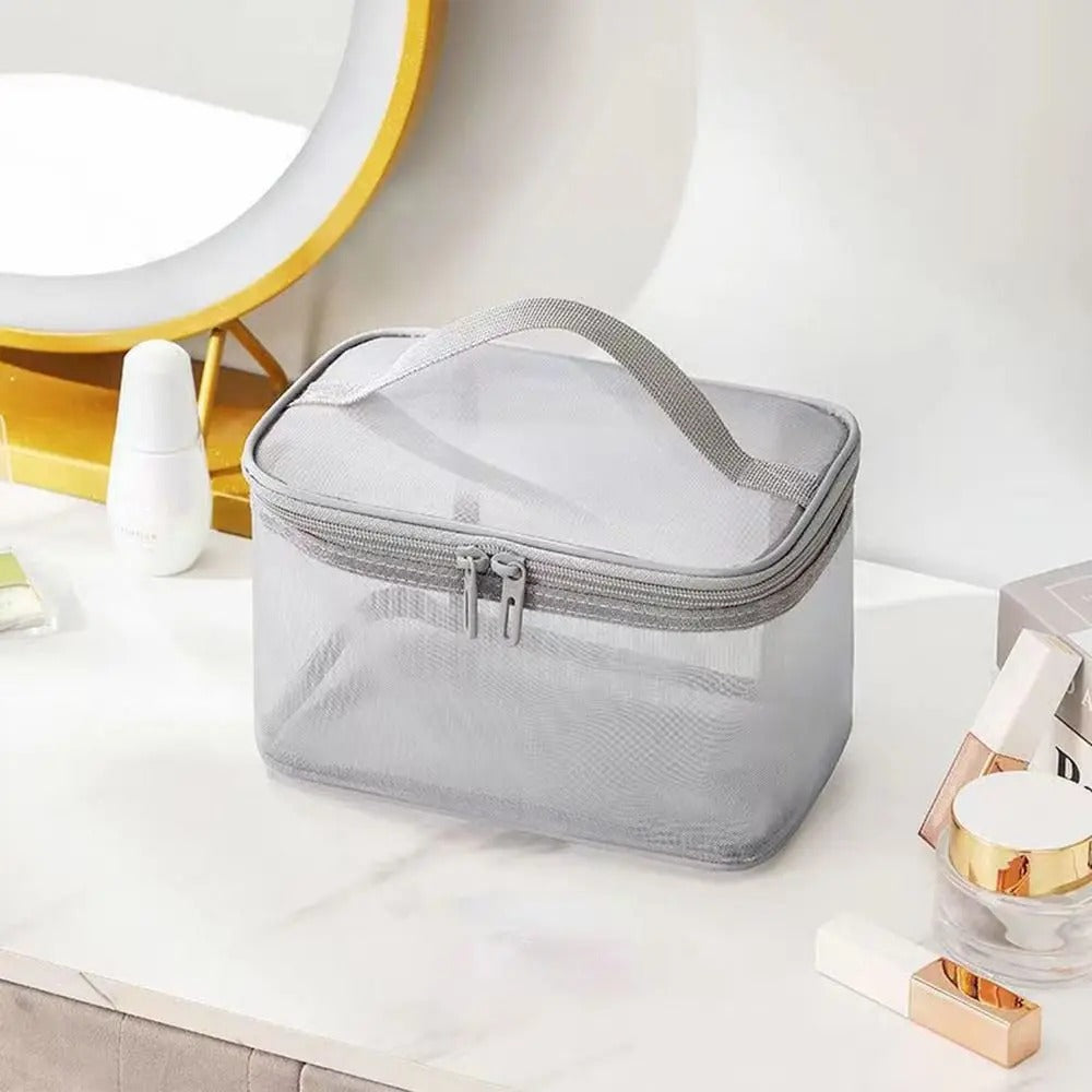 Trousse de toilette femme élégante et transparente, posée sur une coiffeuse blanche.
La trousse de maquillage rectangulaire en maille grise est dotée d'une fermeture éclair double et d'une anse sur le dessus.
Divers produits de beauté tels que des flacons blancs et des tubes dorés l'entourent.
Un miroir rond avec un cadre doré est visible en arrière-plan, créant une ambiance soignée.