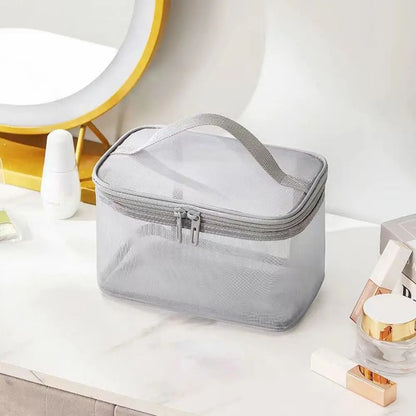 Trousse de toilette femme élégante et transparente, posée sur une coiffeuse blanche.
La trousse de maquillage rectangulaire en maille grise est dotée d'une fermeture éclair double et d'une anse sur le dessus.
Divers produits de beauté tels que des flacons blancs et des tubes dorés l'entourent.
Un miroir rond avec un cadre doré est visible en arrière-plan, créant une ambiance soignée.