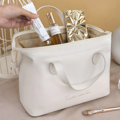 Une main féminine dépose délicatement un tube de crème blanche dans une Trousse de toilette femme luxe. en cuir blanc cassé. La trousse ouverte révèle un intérieur beige soyeux et contient un flacon de parfum doré et transparent ainsi qu'un poudrier compact doré au design géométrique. L'inscription cursive "Twinkle little Star" orne le devant de la trousse, posée sur une surface beige avec un pinceau de maquillage à manche doré à proximité. Trousse de toilette femme luxe.