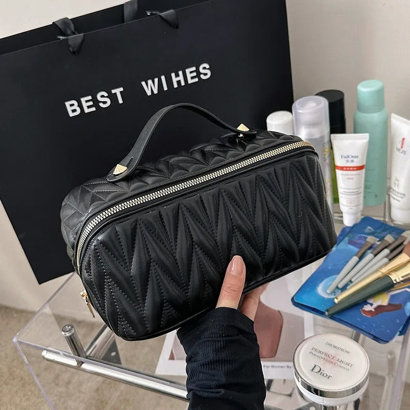 Trousse de toilette femme matelassée noire, élégante et compacte, tenue par une main devant un assortiment de produits de beauté et un sac de courses noir portant l'inscription "BEST WISHES".