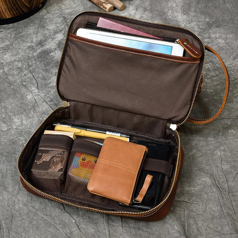 Trousse_de_toilette_homme en cuir ouvert sur une surface grise, contenant un portefeuille, une tablette électronique, deux stylos, une pochette colorée et un petit carnet beige. Le sac comporte plusieurs compartiments, dont des poches en filet et une section zippée.