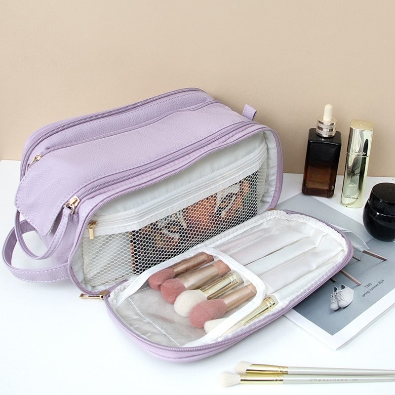 Trousse de toilette luxe femme ouverte en cuir lavande.
Des pinceaux de maquillage aux manches dorés sont rangés dans un compartiment intérieur.
Des flacons de cosmétiques et un pot noir sont disposés à côté.
L'ensemble repose sur une surface blanche avec un arrière-plan beige uni.