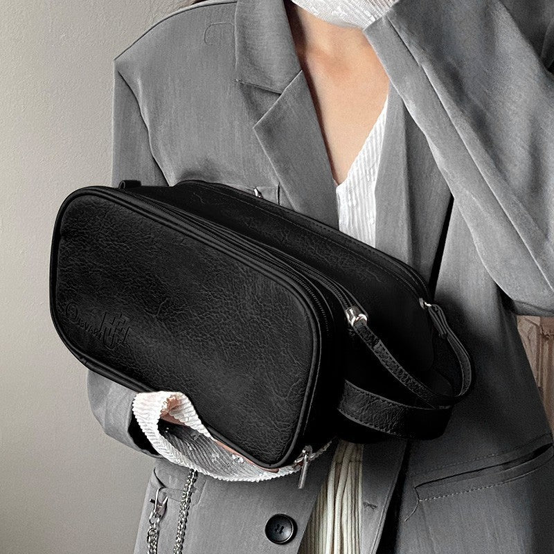 Une femme élégante portant un blazer gris tient une Trousse de toilette luxe femme noire texturée. La trousse a une fermeture éclair argentée et une courte sangle de transport. Un détail de chaîne argentée pend sur le côté. Le nom "Élégance Noire" accompagne le texte "Trousse de toilette luxe femme".