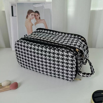 Trousse de toilette luxe femme : une élégante trousse de toilette à motif pied-de-poule noir et blanc est posée sur une surface blanche.
Elle possède plusieurs compartiments zippés pratiques et une poignée latérale.
À l'arrière-plan flou, on aperçoit un livre ou un magazine et un rideau clair.
Quelques accessoires de maquillage sont partiellement visibles au premier plan.