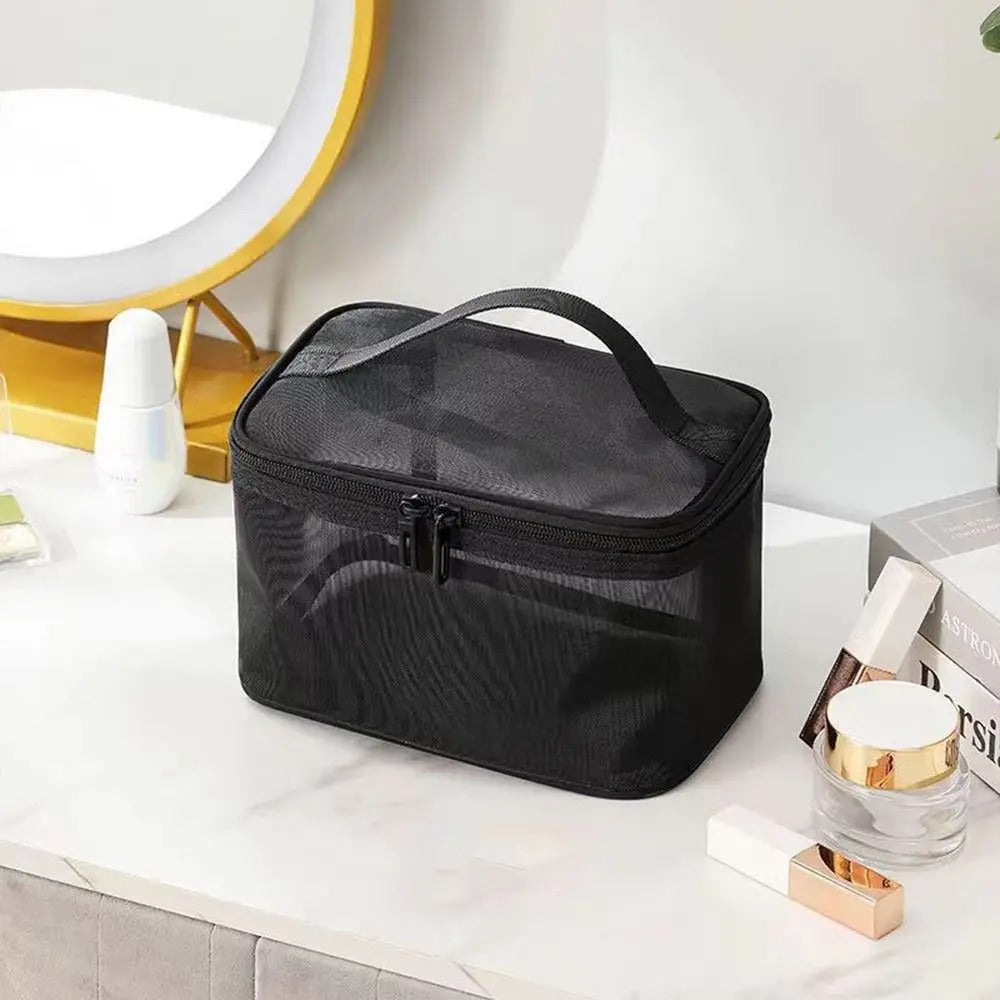 Une Trousse de Toilette Femme noire en maille repose sur une surface marbrée blanche.
Elle est rectangulaire avec une fermeture éclair sur le dessus et une poignée de transport.
Divers produits de beauté, flacons blancs et pots transparents, l'entourent sur la table.
Un miroir rond doré est visible en arrière-plan, suggérant un espace de maquillage.