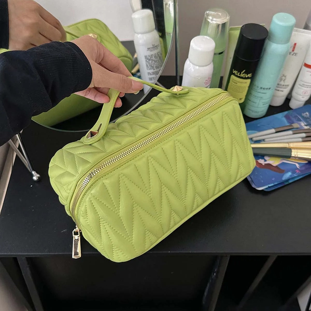 Une Trousse de toilette femme vert vif à motif matelassé en chevrons repose sur une coiffe noire brillante.
Une main gantée de noir soulève le couvercle, révélant un intérieur spacieux.
Divers produits de beauté blancs, verts et noirs sont visibles à l'arrière-plan.
Le reflet d'une autre trousse de toilette verte apparaît dans le miroir. Nom : Élégance en Vert. 