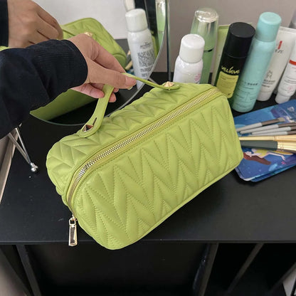 Une Trousse de toilette femme vert vif à motif matelassé en chevrons repose sur une coiffe noire brillante.
Une main gantée de noir soulève le couvercle, révélant un intérieur spacieux.
Divers produits de beauté blancs, verts et noirs sont visibles à l'arrière-plan.
Le reflet d'une autre trousse de toilette verte apparaît dans le miroir. Nom : Élégance en Vert. 