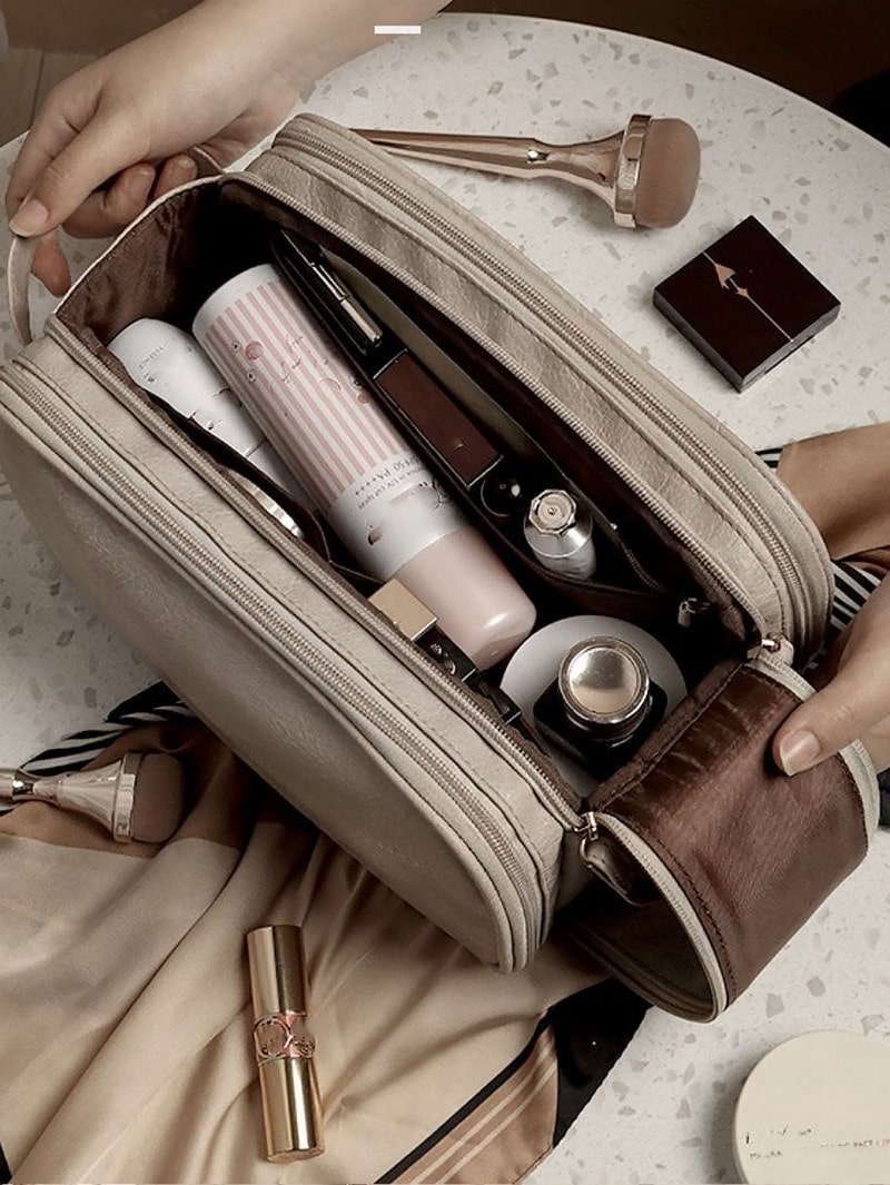 Une Trousse de toilette luxe femme beige et marron, partiellement ouverte, révèle divers produits de beauté. Des mains féminines tiennent la trousse au-dessus d'une surface marbrée claire, entourée d'autres cosmétiques et d'un foulard en soie aux tons doux. Le nom affiché est : Élégance en voyage : Trousse de toilette luxe femme.