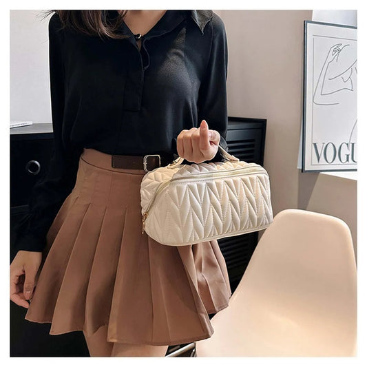 Une femme portant une jupe plissée marron et un chemisier noir tient une petite Trousse de toilette femme blanche matelassée. La trousse a une forme rectangulaire avec une fermeture éclair dorée et une anse sur le dessus. L'arrière-plan est légèrement flou, mais on aperçoit une chaise blanche et un meuble sombre.