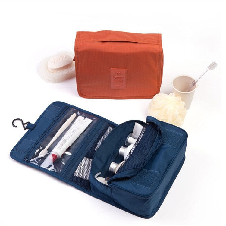 Une trousse de toilette femme luxe, ouverte et de couleur bleu marine, révèle plusieurs compartiments contenant des articles de toilette essentiels tels qu'une brosse à dents, du dentifrice et des flacons de produits. À côté, une trousse de toilette orange, fermée et rectangulaire, repose sur une surface blanche. Un gant de toilette blanc et une petite tasse avec une brosse complètent cet ensemble de voyage élégant.