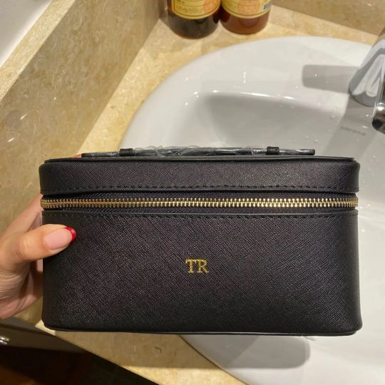 Trousse de maquillage noire personnalisée avec initiales dorées "TR" sur un lavabo clair.