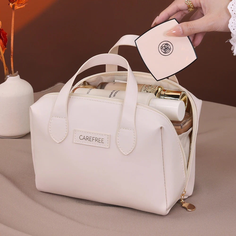 Une Trousse de toilette femme blanche en cuir synthétique repose sur une surface beige. Une main féminine ornée de bagues sort un poudrier beige de la trousse ouverte, révélant divers produits de beauté à l'intérieur. La trousse arbore une étiquette ovale portant l'inscription "CAREFREE".