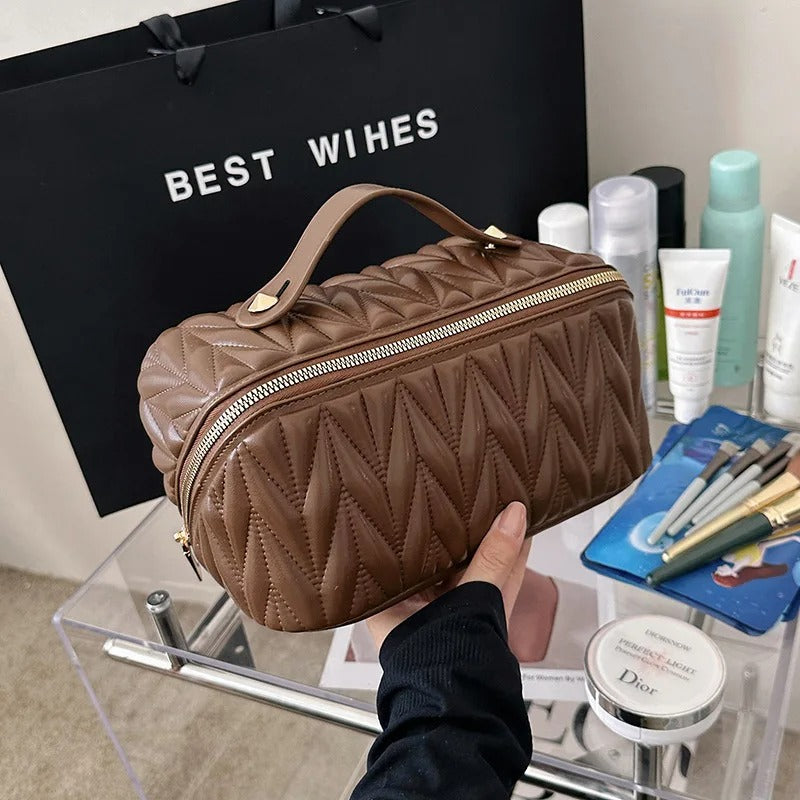 Une trousse de toilette café matelassée, élégante et compacte, est tenue à la main au-dessus d'une table transparente.
Divers produits de beauté, tels que des crèmes, des lotions et des pinceaux de maquillage, sont disposés sur la table à l'arrière-plan.
Un sac cadeau noir portant l'inscription "BEST WISHES" est visible derrière la trousse de toilette.