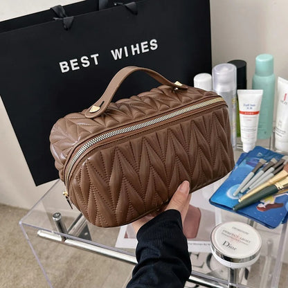 Une trousse de toilette café matelassée, élégante et compacte, est tenue à la main au-dessus d'une table transparente.
Divers produits de beauté, tels que des crèmes, des lotions et des pinceaux de maquillage, sont disposés sur la table à l'arrière-plan.
Un sac cadeau noir portant l'inscription "BEST WISHES" est visible derrière la trousse de toilette.