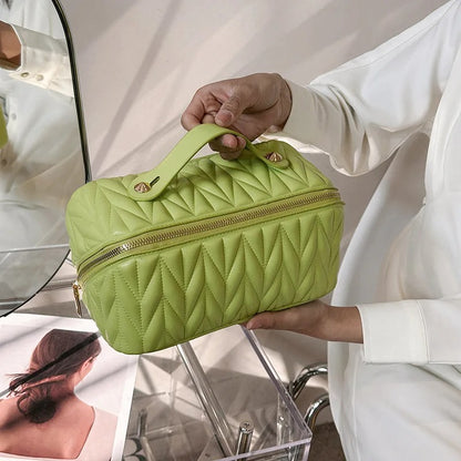 Une trousse de toilette femme en cuir végétal vert vif, ornée de motifs géométriques en relief.
La trousse de forme rectangulaire présente une fermeture éclair dorée sur le dessus et des côtés, ainsi qu'une anse rigide de la même couleur.
Elle est délicatement tenue par deux mains féminines soignées, vêtues d'un chemisier blanc.
Le fond est légèrement flou, suggérant un intérieur clair et minimaliste.