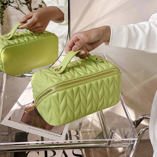 Une main délicate tient une élégante Trousse_de_toilette_femme vert clair, ornée de motifs matelassés en chevrons et de détails dorés.
Posée sur une table transparente, la trousse projette son reflet dans un miroir, créant une double présence visuelle.
Un magazine de mode se trouve sous la trousse, suggérant un univers de beauté et de style.
Le design compact et sophistiqué de cet accessoire en fait une parfaite trousse de toilette femme.