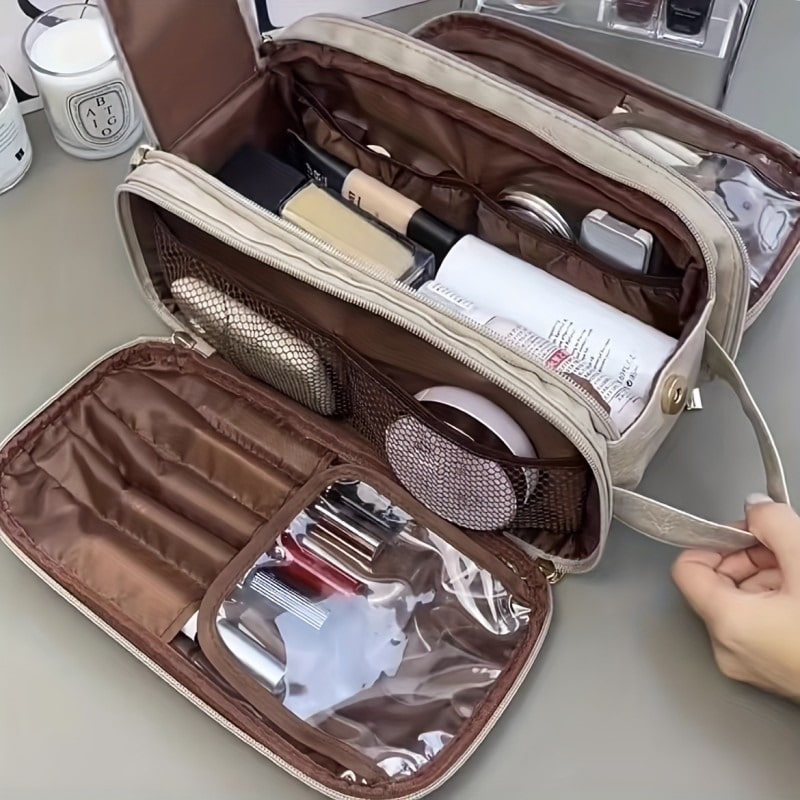 Une trousse de toilette de luxe femme, ouverte et remplie de divers produits de beauté et de maquillage, est posée sur une surface grise. L'intérieur de la trousse est marron et comporte plusieurs compartiments et pochettes pour organiser les articles. Une main féminine tient la poignée de la trousse, suggérant son utilisation ou son déplacement. Des bougies et des flacons de parfum sont visibles en arrière-plan, ajoutant une ambiance de soin et de beauté.
