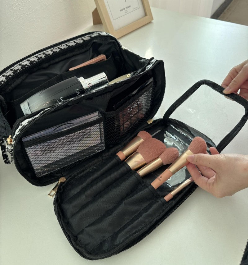 Une trousse de toilette luxe femme est ouverte sur une surface claire. L'intérieur noir et blanc à motif pied-de-poule révèle des compartiments organisés contenant des cosmétiques et des pinceaux de maquillage. Une main féminine ouvre le compartiment transparent zippé contenant des pinceaux aux poils rose pâle.