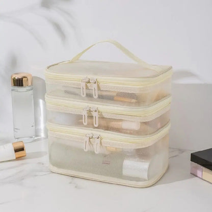Une trousse de toilette femme transparente à trois compartiments repose sur une surface marbrée claire.
La trousse, de couleur crème avec des fermetures éclair dorées, est partiellement remplie de divers produits de beauté.
À gauche, un flacon d'eau micellaire et un tube de crème sont visibles.
À droite, une palette de maquillage complète la scène lumineuse et organisée.