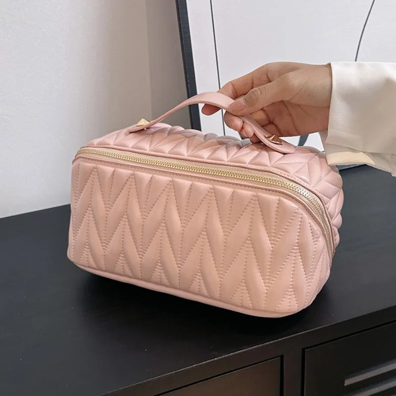 Une main tenant une élégante Trousse de toilette femme rose pâle, matelassée avec un motif de losanges.
La trousse, de forme rectangulaire aux coins arrondis, possède une fermeture éclair dorée sur le dessus.
Un petit manche permet de la porter facilement.