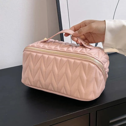 Une main tenant une élégante Trousse de toilette femme rose pâle, matelassée avec un motif de losanges.
La trousse, de forme rectangulaire aux coins arrondis, possède une fermeture éclair dorée sur le dessus.
Un petit manche permet de la porter facilement.