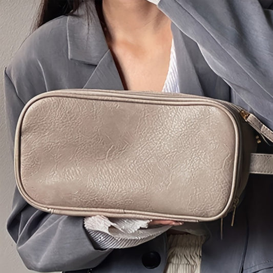 Une femme élégante tient une Trousse de toilette luxe femmerectangulaire en cuir texturé beige clair. La trousse est zippée sur le dessus et a une petite lanière sur le côté. La femme porte un chemisier blanc, une veste grise et ses mains aux ongles soignés sont visibles. Le fond est un mur gris uni.