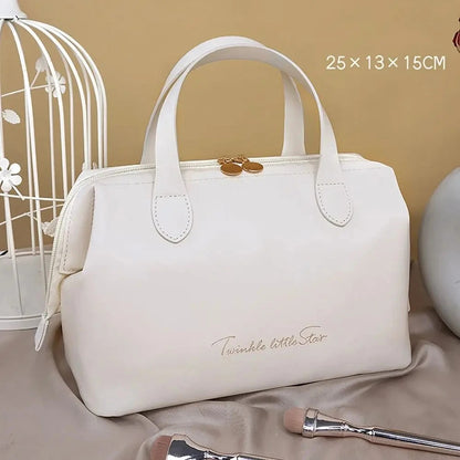 Élégante Trousse de toilette femme luxe blanche en cuir synthétique, de forme rectangulaire avec des coins arrondis et deux anses sur le dessus. Une fermeture éclair dorée sécurise le contenu. L'inscription délicate "Twinkle little Star" orne la face avant en lettres cursives dorées. La trousse est posée sur un tissu satiné beige, avec un coin de cage décorative blanche visible à gauche et des pinceaux de maquillage au premier plan.