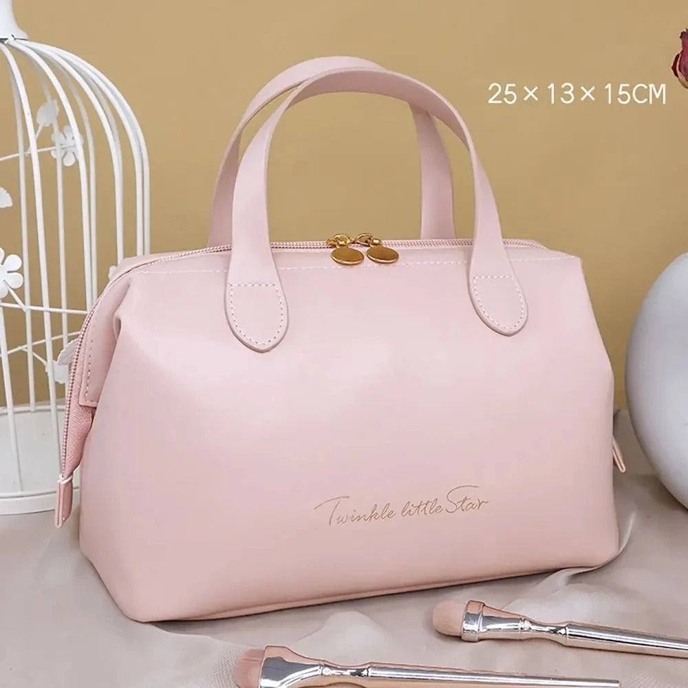 Élégante Trousse de toilette femme luxe Rose en cuir synthétique, de forme rectangulaire avec des coins arrondis et deux anses sur le dessus. Une fermeture éclair dorée sécurise le contenu. L'inscription délicate "Twinkle little Star" orne la face avant en lettres cursives dorées. La trousse est posée sur un tissu satiné beige, avec un coin de cage décorative blanche visible à gauche et des pinceaux de maquillage au premier plan.
