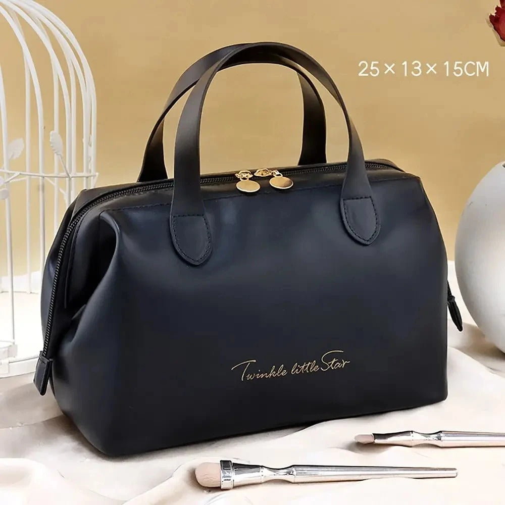 Élégante Trousse de toilette femme luxe noir en cuir synthétique, de forme rectangulaire avec des coins arrondis et deux anses sur le dessus. Une fermeture éclair dorée sécurise le contenu. L'inscription délicate "Twinkle little Star" orne la face avant en lettres cursives dorées. La trousse est posée sur un tissu satiné beige, avec un coin de cage décorative blanche visible à gauche et des pinceaux de maquillage au premier plan.