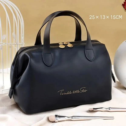 Élégante Trousse de toilette femme luxe noir en cuir synthétique, de forme rectangulaire avec des coins arrondis et deux anses sur le dessus. Une fermeture éclair dorée sécurise le contenu. L'inscription délicate "Twinkle little Star" orne la face avant en lettres cursives dorées. La trousse est posée sur un tissu satiné beige, avec un coin de cage décorative blanche visible à gauche et des pinceaux de maquillage au premier plan.