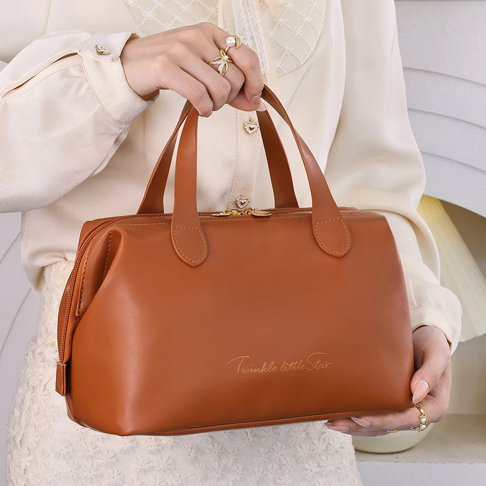 Une femme aux mains délicatement parées de bagues tient un élégant sac de toilette en cuir marron clair. La trousse de toilette a une forme structurée et est ornée d'une discrète inscription dorée "Twinkle little Star". Les finitions dorées de la fermeture éclair et des attaches des anses ajoutent une touche de luxe. Le fond crème et le vêtement texturé de la femme mettent en valeur l'accessoire de voyage raffiné, intitulé