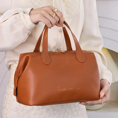 Une femme aux mains délicatement parées de bagues tient un élégant sac de toilette en cuir marron clair. La trousse de toilette a une forme structurée et est ornée d'une discrète inscription dorée "Twinkle little Star". Les finitions dorées de la fermeture éclair et des attaches des anses ajoutent une touche de luxe. Le fond crème et le vêtement texturé de la femme mettent en valeur l'accessoire de voyage raffiné, intitulé