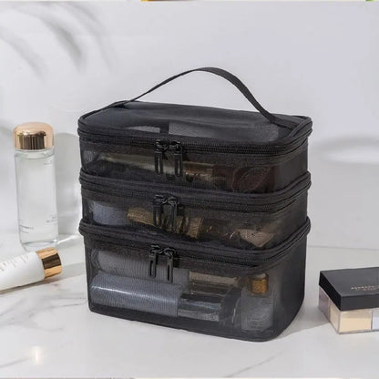 Une trousse-de-toilette-femme noire à trois compartiments en maille repose sur une surface en marbre blanc. Chaque compartiment est fermé par une fermeture éclair noire à double curseur. Une poignée noire est fixée sur le dessus de la trousse. À gauche, on aperçoit partiellement des flacons de produits de beauté, et à droite, une boîte noire et dorée.