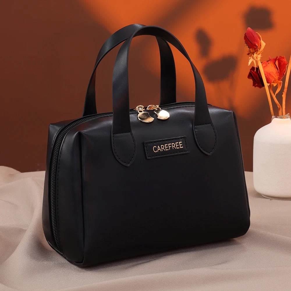 Trousse de toilette femme" sur une étiquette apposée sur une élégante trousse de toilette Noir, de forme structurée avec des anses, posée sur un tissu clair et froissé, avec un fond brun et un vase blanche contenant des fleurs séchées rouges. Le nom de la trousse sera "Carefree".