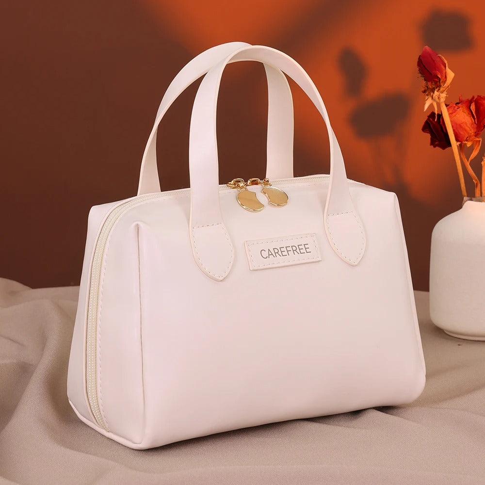 Trousse de toilette femme" sur une étiquette apposée sur une élégante trousse de toilette blanche, de forme structurée avec des anses, posée sur un tissu clair et froissé, avec un fond brun et un vase blanc contenant des fleurs séchées rouges. Le nom de la trousse sera "Carefree".