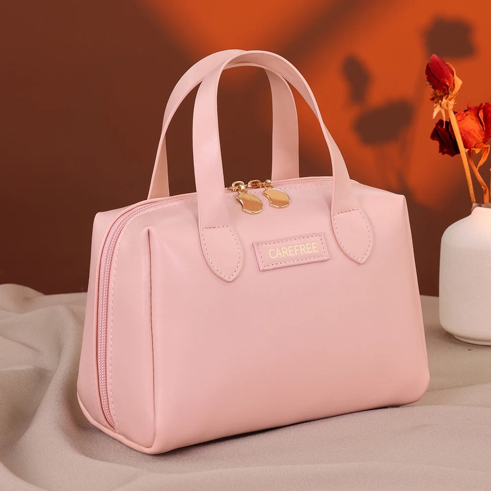 Trousse de toilette femme" sur une étiquette apposée sur une élégante trousse de toilette rose, de forme structurée avec des anses, posée sur un tissu clair et froissé, avec un fond brun et un vase blanche contenant des fleurs séchées rouges. Le nom de la trousse sera "Carefree".