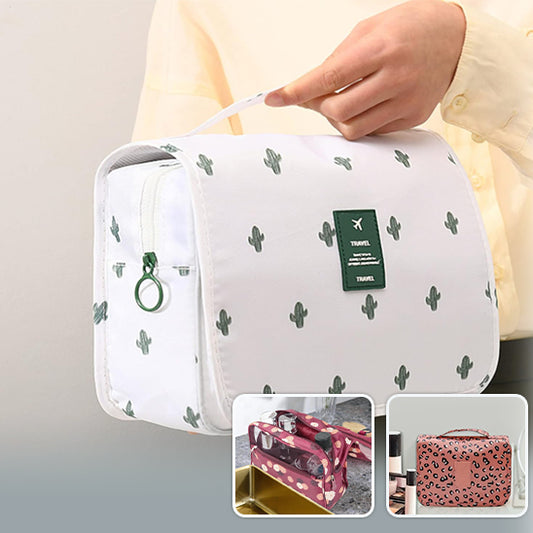 Une main tient une élégante trousse de toilette femme luxe blanche ornée de motifs de cactus verts. La trousse de toilette de luxe, idéale pour les voyages, présente une fermeture éclair verte et une étiquette verte portant le mot "TRAVEL". En arrière-plan, on aperçoit d'autres trousses, dont une à motifs de coeurs et une à imprimé léopard, suggérant une variété de styles pour la femme moderne.